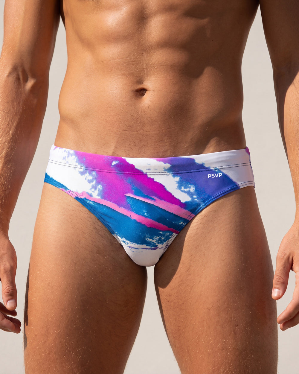 Close-up front view of a male model wearing a speedo-style swim brief with a 90s-Jazz-inspired pattern (white, blue, pink, and purple) on a white background by PARADIS SVP