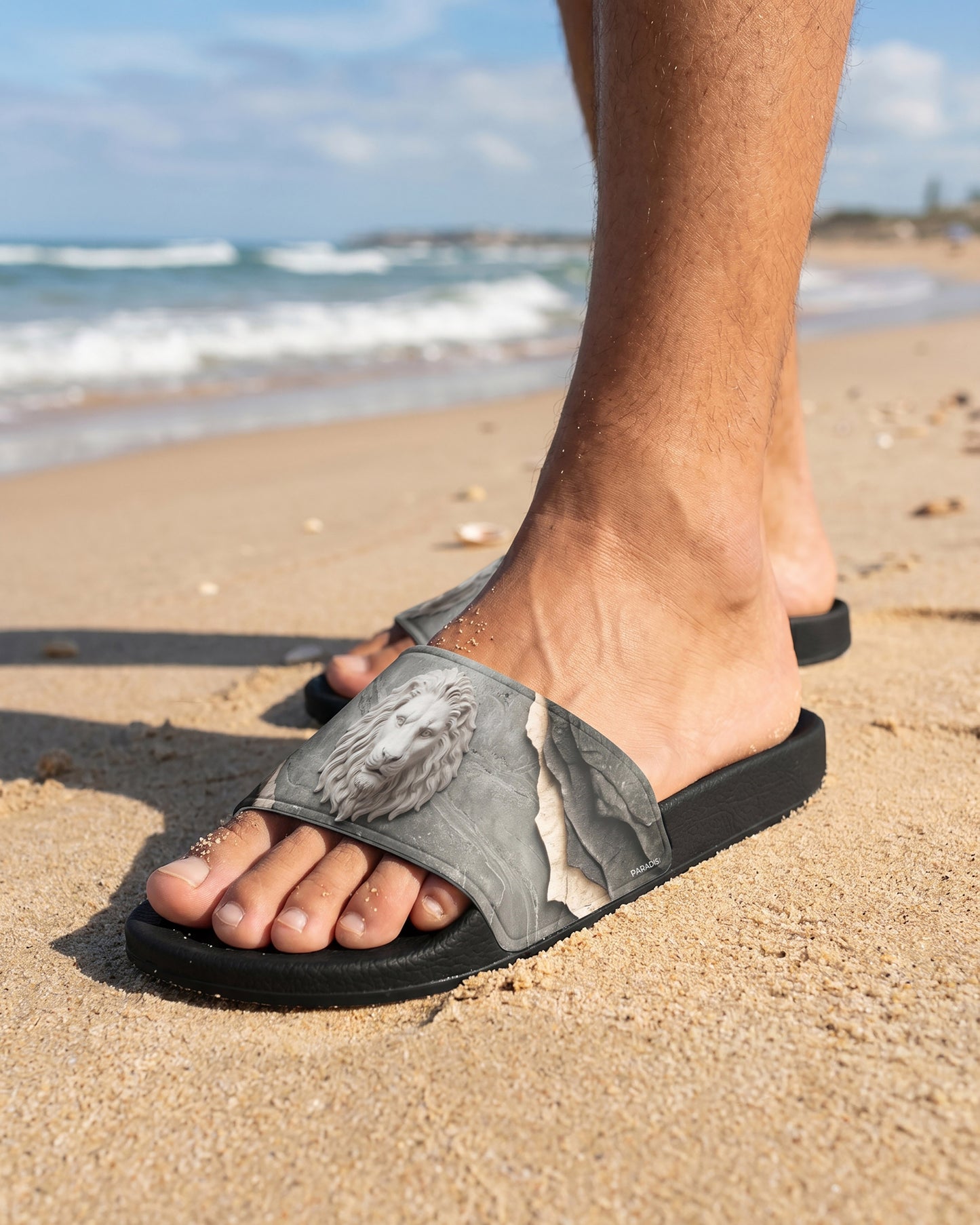 Male model wearing a grey and black Pair of slides with a pattern of a lion on a sandy beach