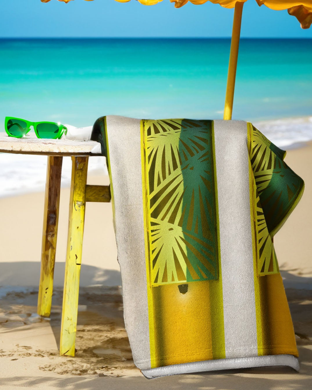 Beach towel with green palm leaf design on a yellow and white striped background, placed on sand with ocean view.
