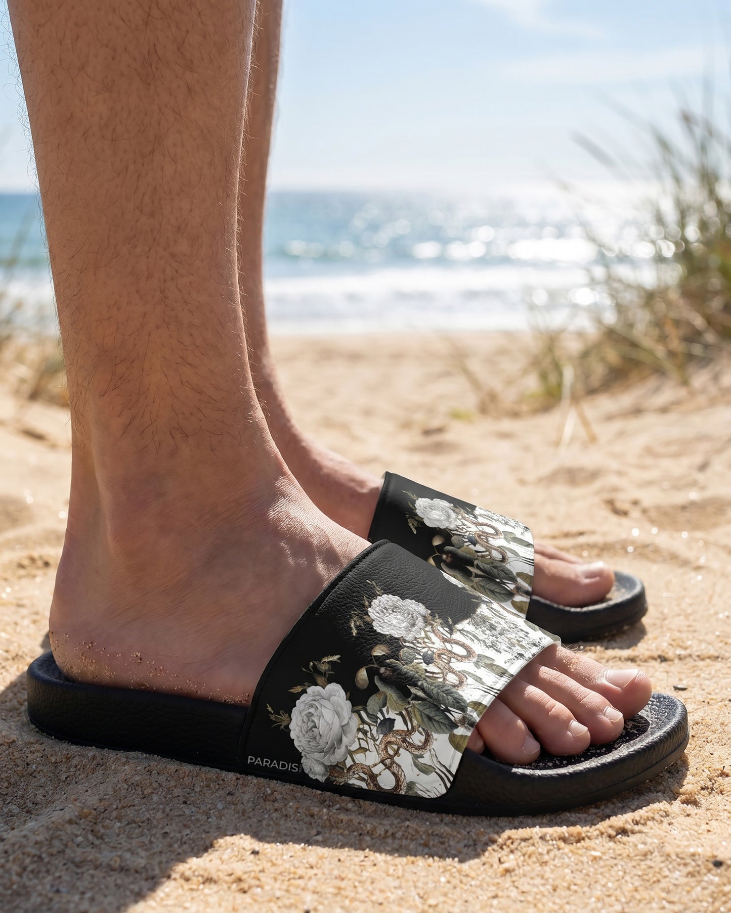 Person wearing floral slides on a sandy beach with ocean view